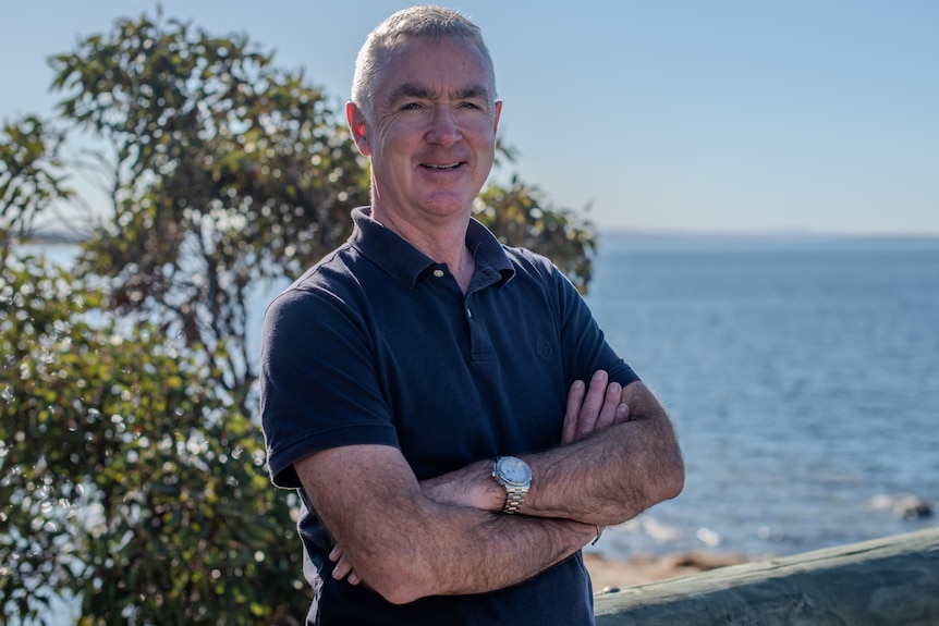 A man crosses his arms and smiles, with the ocean in the background.